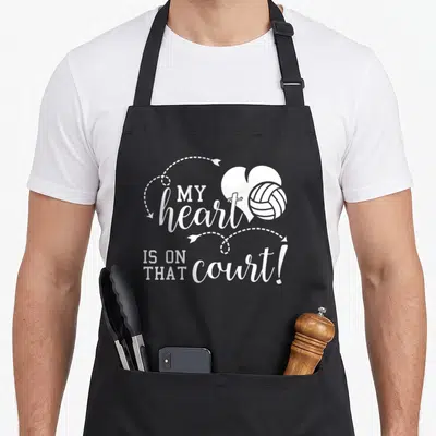 Volleyball school dad apron - White typography reading MY heart IS ON THAT court with a heart and volleyball graphic — shown on a black cotton canvas apron against a white background