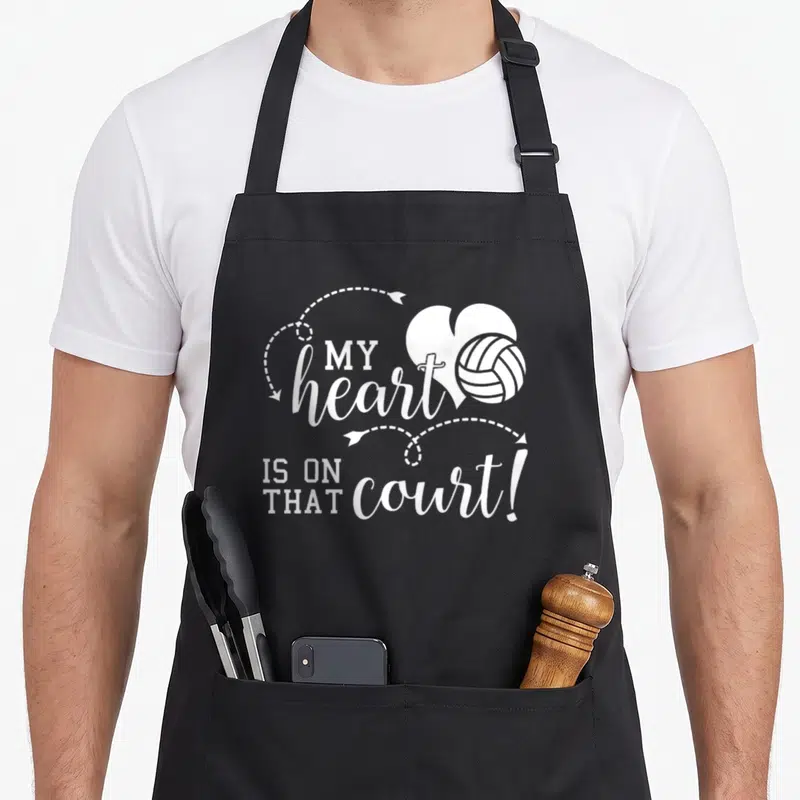 Volleyball school dad apron - White typography reading MY heart IS ON THAT court with a heart and volleyball graphic — shown on a black cotton canvas apron against a white background