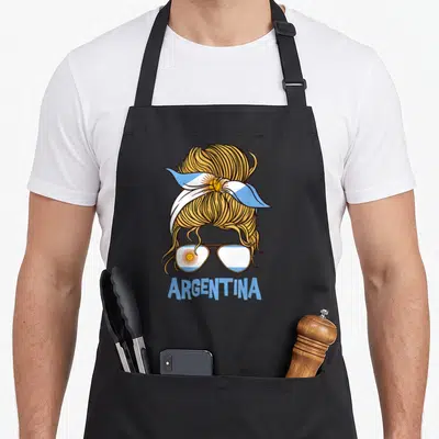 Argentinian argentine apron - Blonde messy bun with flag headband and sunglasses below ARGENTINA text — shown on a black cotton canvas apron against a white background
