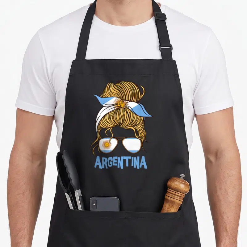 Argentinian argentine apron - Blonde messy bun with flag headband and sunglasses below ARGENTINA text — shown on a black cotton canvas apron against a white background