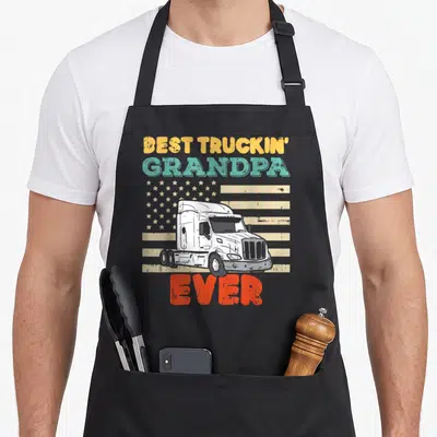 Truckin american flag apron - Semi-truck over distressed flag with Best Truckin Grandpa Ever text — shown on a black cotton canvas apron against a white background
