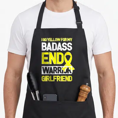 Girlfriend endometriosis awareness apron - Stacked yellow and white text I GO YELLOW FOR MY BADASS ENDO WARRIOR — shown on a black cotton canvas apron against a white background