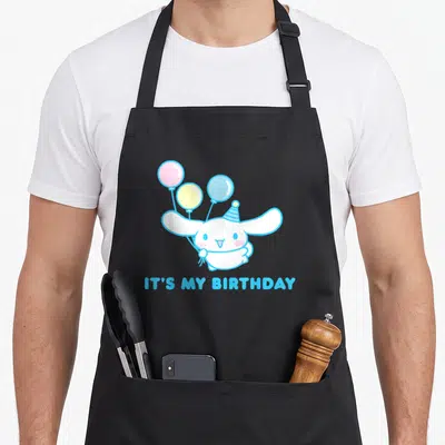 Birthday cinnamoroll apron - White creature holding three balloons with IT'S MY BIRTHDAY text in blue — shown on a black cotton canvas apron against a white background