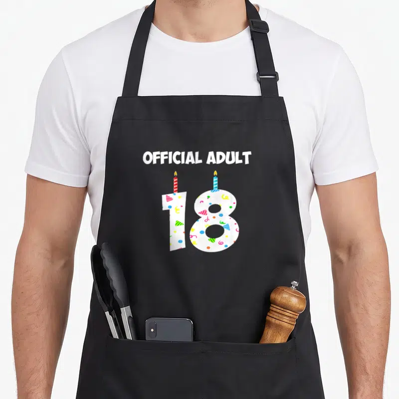 Candle birthday apron - OFFICIAL ADULT text above white number 18 with two candles and confetti — shown on a black cotton canvas apron against a white background