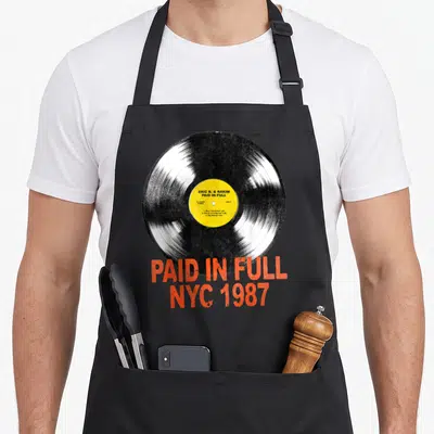 Eric vinyl apron - Black vinyl record with yellow label and distressed orange-red Paid In Full NYC 1987 text — shown on a black cotton canvas apron against a white background