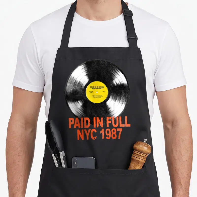 Eric vinyl apron - Black vinyl record with yellow label and distressed orange-red Paid In Full NYC 1987 text — shown on a black cotton canvas apron against a white background