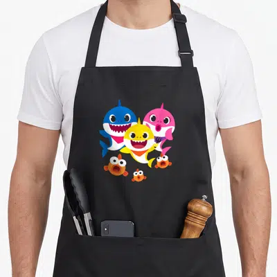 Shark family aprons - Three cartoon sharks in blue pink yellow with small orange fish below — shown on a black cotton canvas apron against a white background