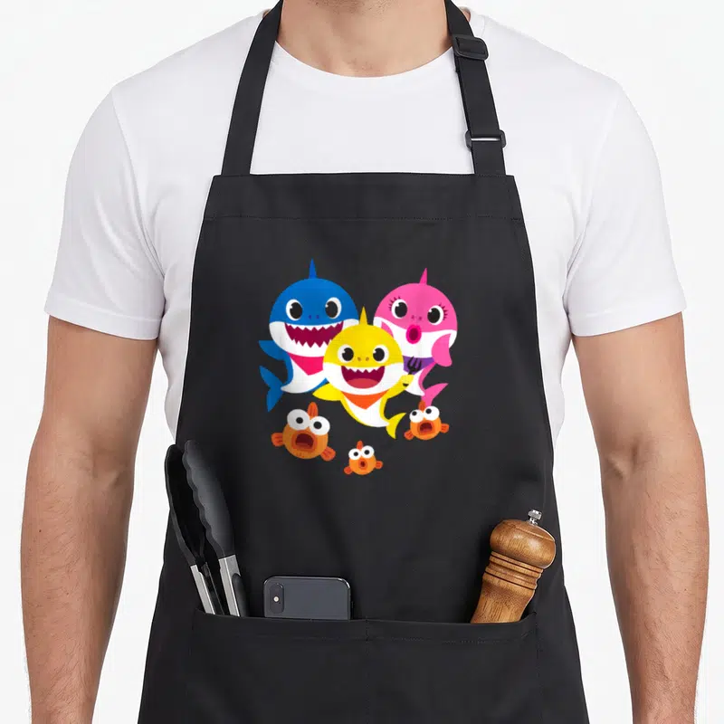 Shark family aprons - Three cartoon sharks in blue pink yellow with small orange fish below — shown on a black cotton canvas apron against a white background