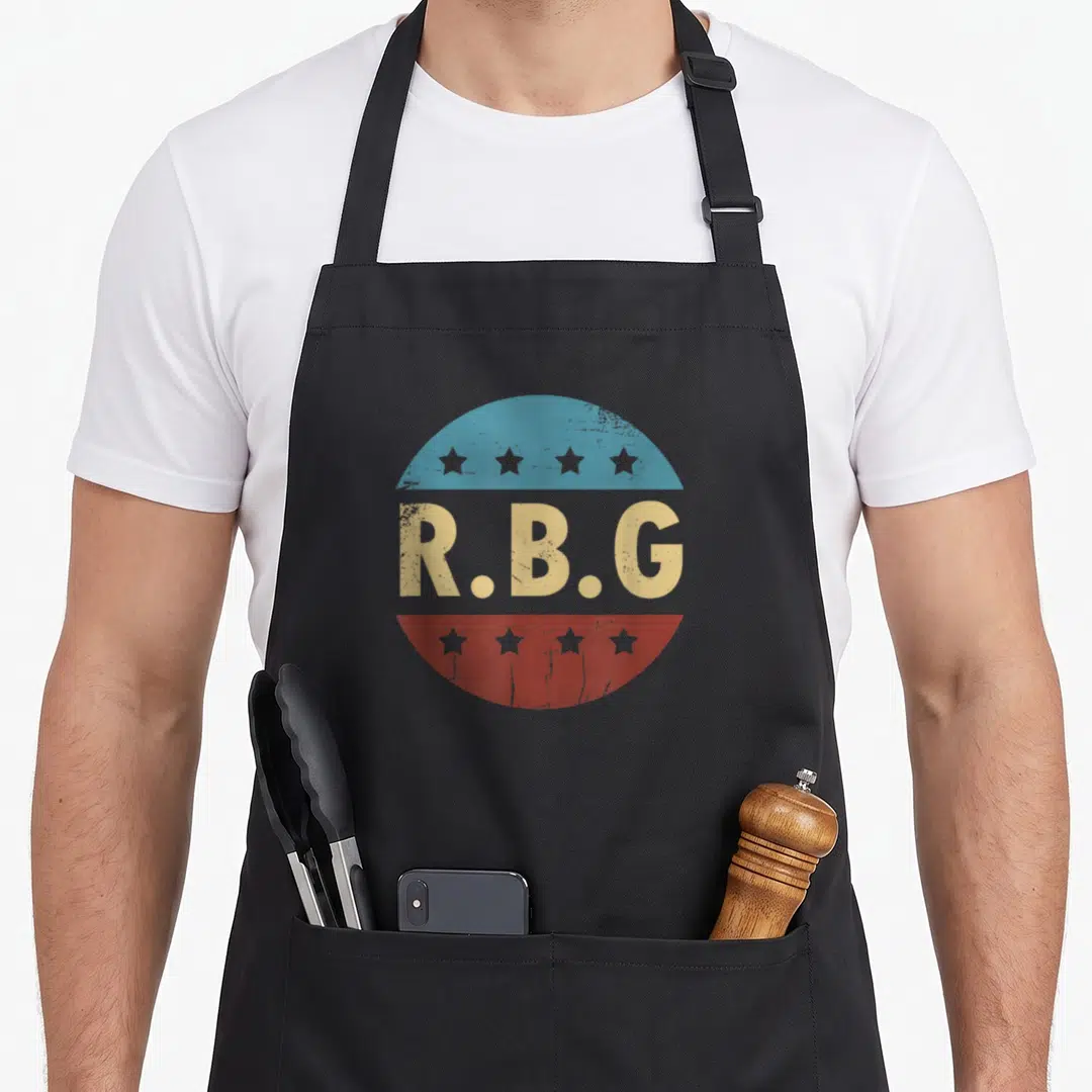 Rbg tribute apron - R.B.G text in distressed beige over blue and red circle with eight stars total — shown on a black cotton canvas apron against a white background