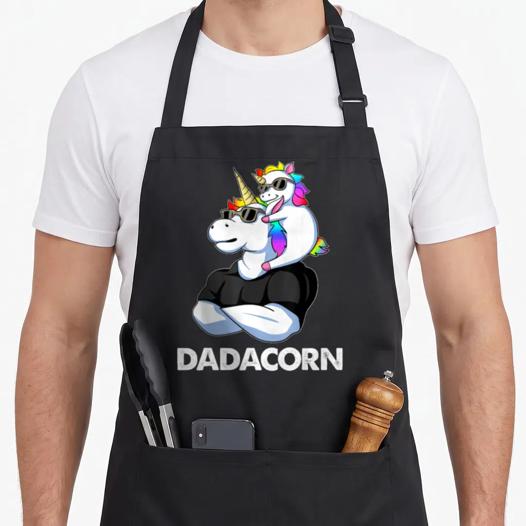 Dadacorn papa apron - Cartoon unicorn dad in black shirt with sunglasses carries baby unicorn; DADACORN text below — shown on a black cotton canvas apron against a white background