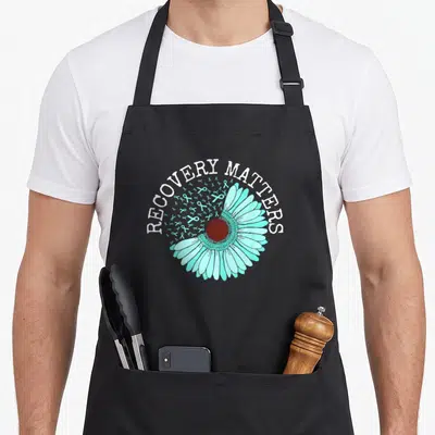 Recovery ribbon apron - RECOVERY MATTERS text over turquoise daisy and scattered ribbons — shown on a black cotton canvas apron against a white background