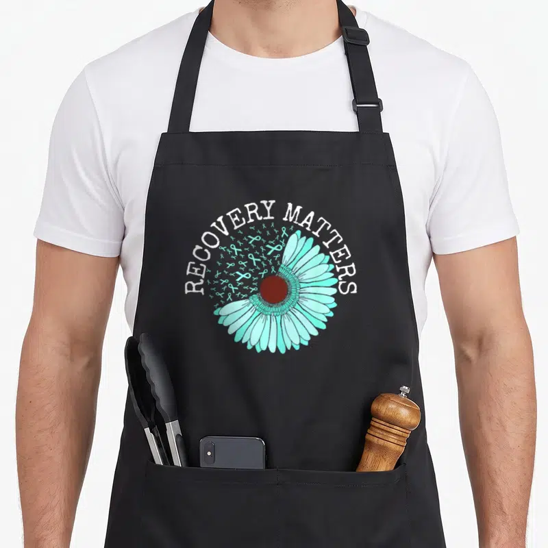 Recovery ribbon apron - RECOVERY MATTERS text over turquoise daisy and scattered ribbons — shown on a black cotton canvas apron against a white background