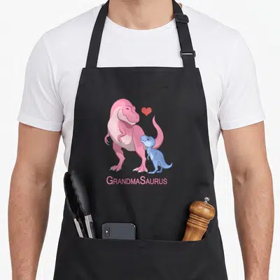 Grandmasaurus dinosaur apron - Pink T-Rex and blue baby dinosaur with red heart and GRANDMASAURUS text — shown on a black cotton canvas apron against a white background