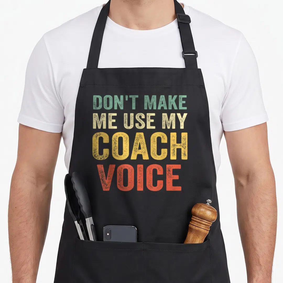 Coach use of apron - Stacked text DON'T MAKE ME USE MY COACH VOICE in green beige yellow orange distressed font — shown on a black cotton canvas apron against a white background