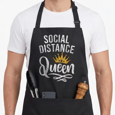 Social at home aprons - White distressed text SOCIAL DISTANCE Queen with yellow crown and decorative swirl — shown on a black cotton canvas apron against a white background