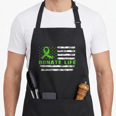 Flag donate organ apron - Distressed green ribbon and white stripes with DONATE LIFE text on a flag background — shown on a black cotton canvas apron against a white background