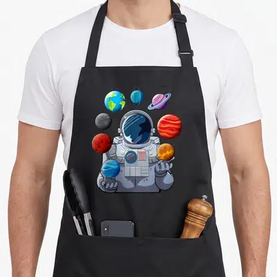 Astronaut space planets apron - Cartoon astronaut holding colorful planets in grey blue red and purple — shown on a black cotton canvas apron against a white background