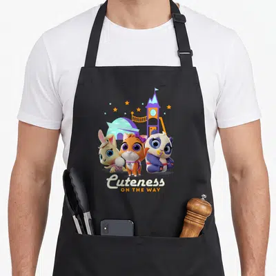 Cuteness junior apron - Three baby animals and stars above playground with Cuteness ON THE WAY text — shown on a black cotton canvas apron against a white background