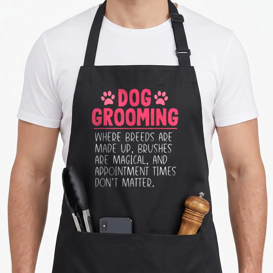 Groomer top dog aprons - Pink DOG GROOMING text flanked by paw prints above white sarcastic quote about breeds — shown on a black cotton canvas apron against a white background