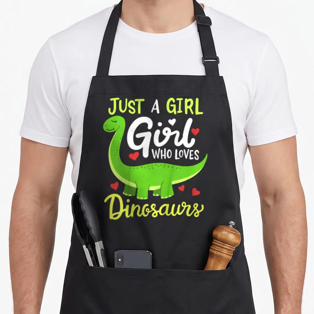 Brachiosaurus dinosaur apron - Green cartoon brachiosaurus with red hearts and text JUST A GIRL Girl WHO LOVES Dinosaurs — shown on a black cotton canvas apron against a white background