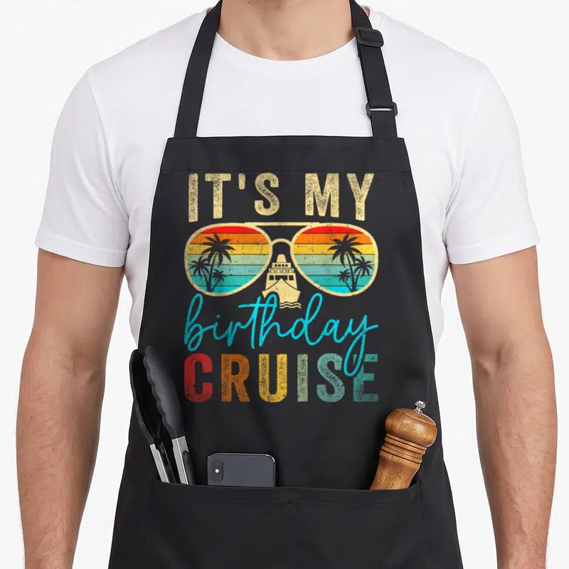 Birthday cruise family aprons - Retro sunglasses containing a sunset, palm trees, and cruise ship with stacked text — shown on a black cotton canvas apron against a white background