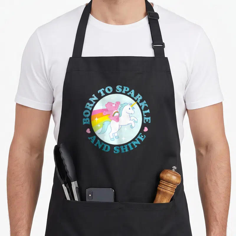 Sparkle care apron - Circular design with pink bear riding white unicorn surrounded by blue text and two pink hearts — shown on a black cotton canvas apron against a white background