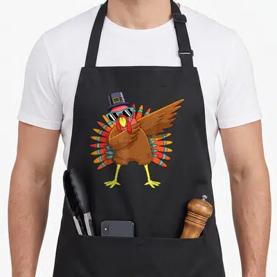 Dab pilgrim apron - Cartoon brown turkey wearing blue hat and sunglasses in a dab pose — shown on a black cotton canvas apron against a white background