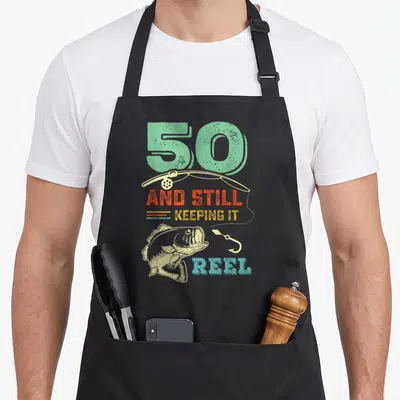 50th fishing aprons - Retro fish and rod with '50 AND STILL KEEPING IT REEL' in mint orange yellow blue beige — shown on a black cotton canvas apron against a white background