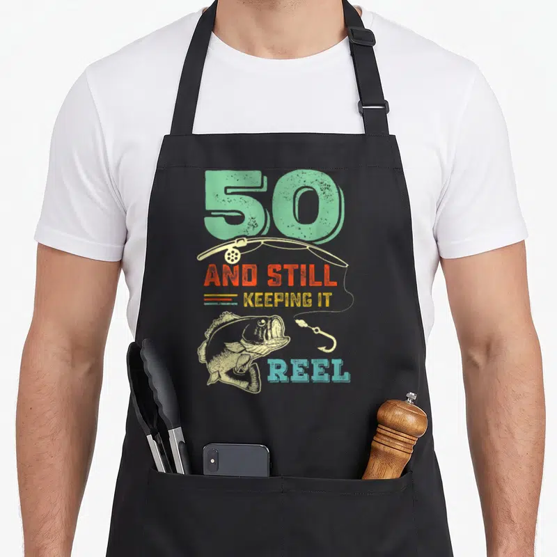 50th fishing aprons - Retro fish and rod with '50 AND STILL KEEPING IT REEL' in mint orange yellow blue beige — shown on a black cotton canvas apron against a white background