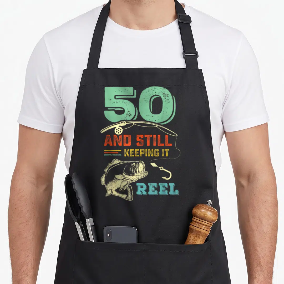 50th fishing aprons - Retro fish and rod with '50 AND STILL KEEPING IT REEL' in mint orange yellow blue beige — shown on a black cotton canvas apron against a white background