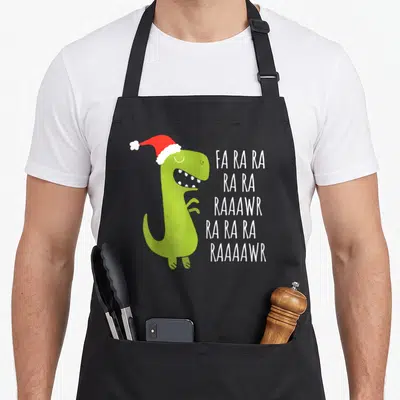 Rawr t-rex apron - Green dinosaur in red Santa hat next to white FA RA RA RAAAWR text — shown on a black cotton canvas apron against a white background