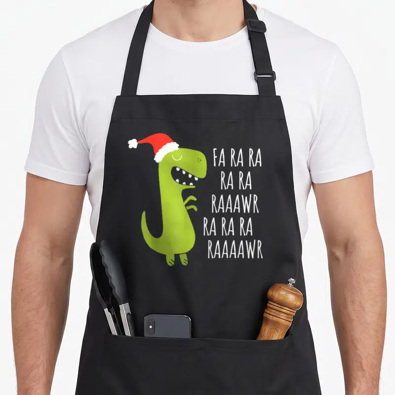 Rawr t-rex apron - Green dinosaur in red Santa hat next to white FA RA RA RAAAWR text — shown on a black cotton canvas apron against a white background