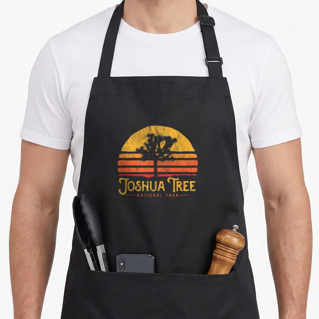 National tree apron - Retro striped sun with yellow orange red colors and dark grey Joshua tree silhouette above — shown on a black cotton canvas apron against a white background