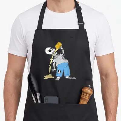 Homer skeleton apron - Cartoon skeleton in grey shirt and blue pants pouring yellow liquid into open mouth — shown on a black cotton canvas apron against a white background