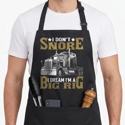 Snore truck apron - Distressed white semi-truck with tan text reading I DON'T SNORE I DREAM I'M A BIG RIG flanked — shown on a black cotton canvas apron against a white background