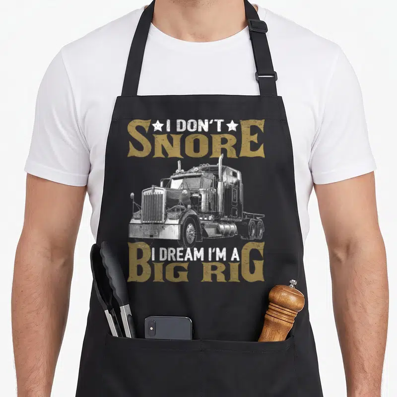 Snore truck apron - Distressed white semi-truck with tan text reading I DON'T SNORE I DREAM I'M A BIG RIG flanked — shown on a black cotton canvas apron against a white background