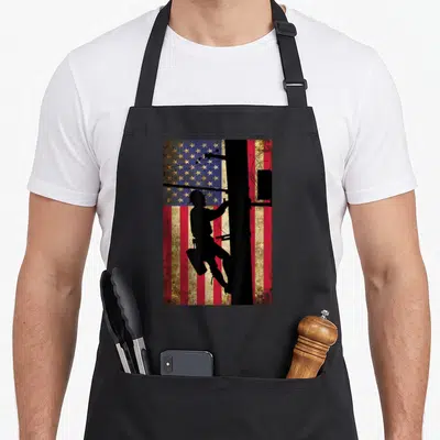 Lineman american flag apron - Black lineman silhouette climbing pole over distressed red white and blue flag — shown on a black cotton canvas apron against a white background