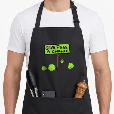 Chance peas apron - Lime green sign reading GIVE PEAS A CHANCE with four cartoon peas at the base of a brown post — shown on a black cotton canvas apron against a white background