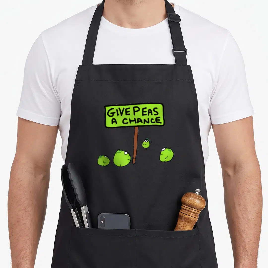 Chance peas apron - Lime green sign reading GIVE PEAS A CHANCE with four cartoon peas at the base of a brown post — shown on a black cotton canvas apron against a white background