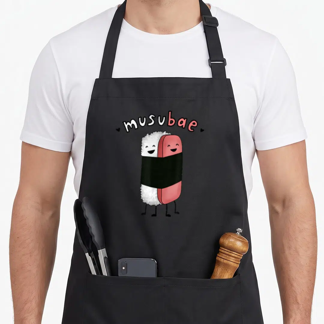 Musubi hawaiian apron - Two cartoon onigiri wrapped in black seaweed under arched text reading musubae with small hearts — shown on a black cotton canvas apron against a white background