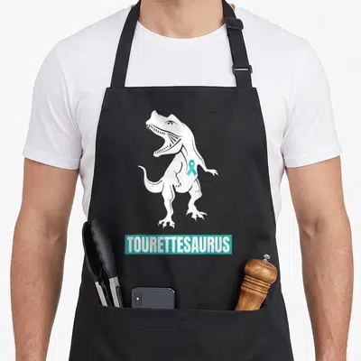 Tourette ribbon apron - White dinosaur silhouette with teal ribbon and TOURETTESAURUS text in a teal bar — shown on a black cotton canvas apron against a white background