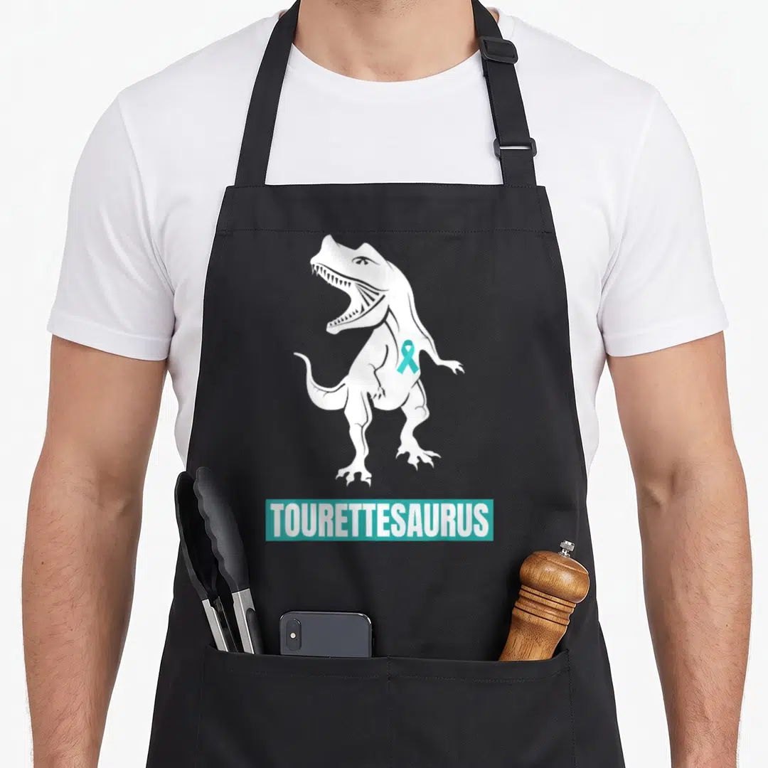 Tourette ribbon apron - White dinosaur silhouette with teal ribbon and TOURETTESAURUS text in a teal bar — shown on a black cotton canvas apron against a white background
