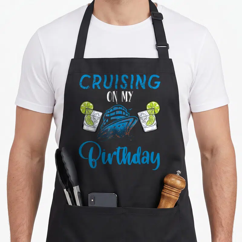 Boarding birthday apron - Cruising On My Birthday text with blue ship and two lime cocktails — shown on a black cotton canvas apron against a white background