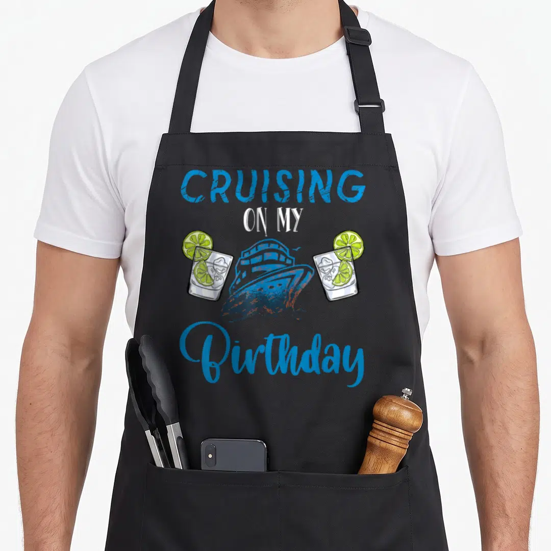 Boarding birthday apron - Cruising On My Birthday text with blue ship and two lime cocktails — shown on a black cotton canvas apron against a white background