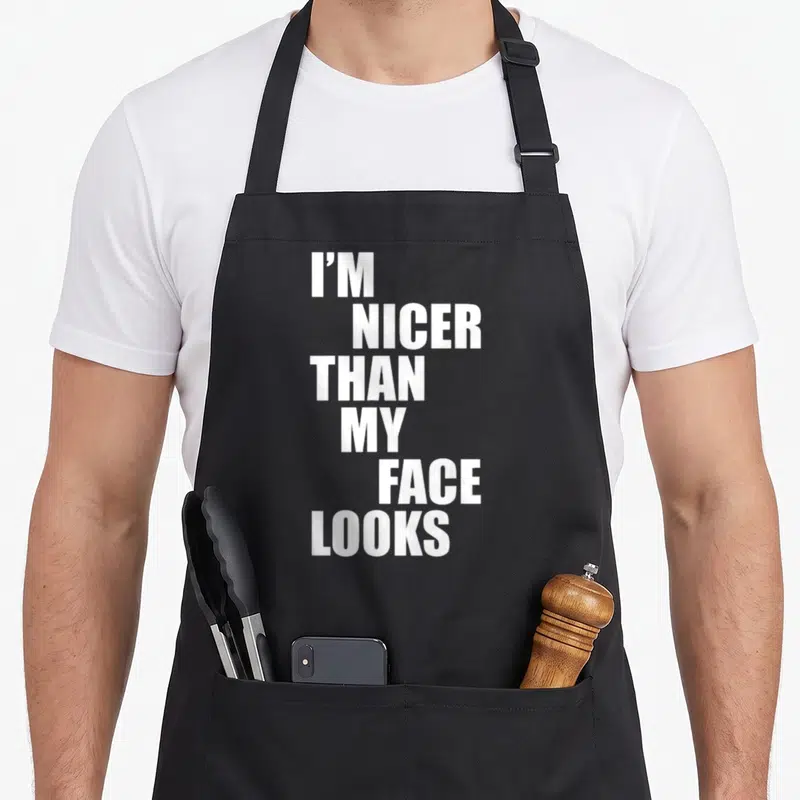 Nicer face apron - White stacked text reading I'M NICER THAN MY FACE LOOKS on dark background — shown on a black cotton canvas apron against a white background