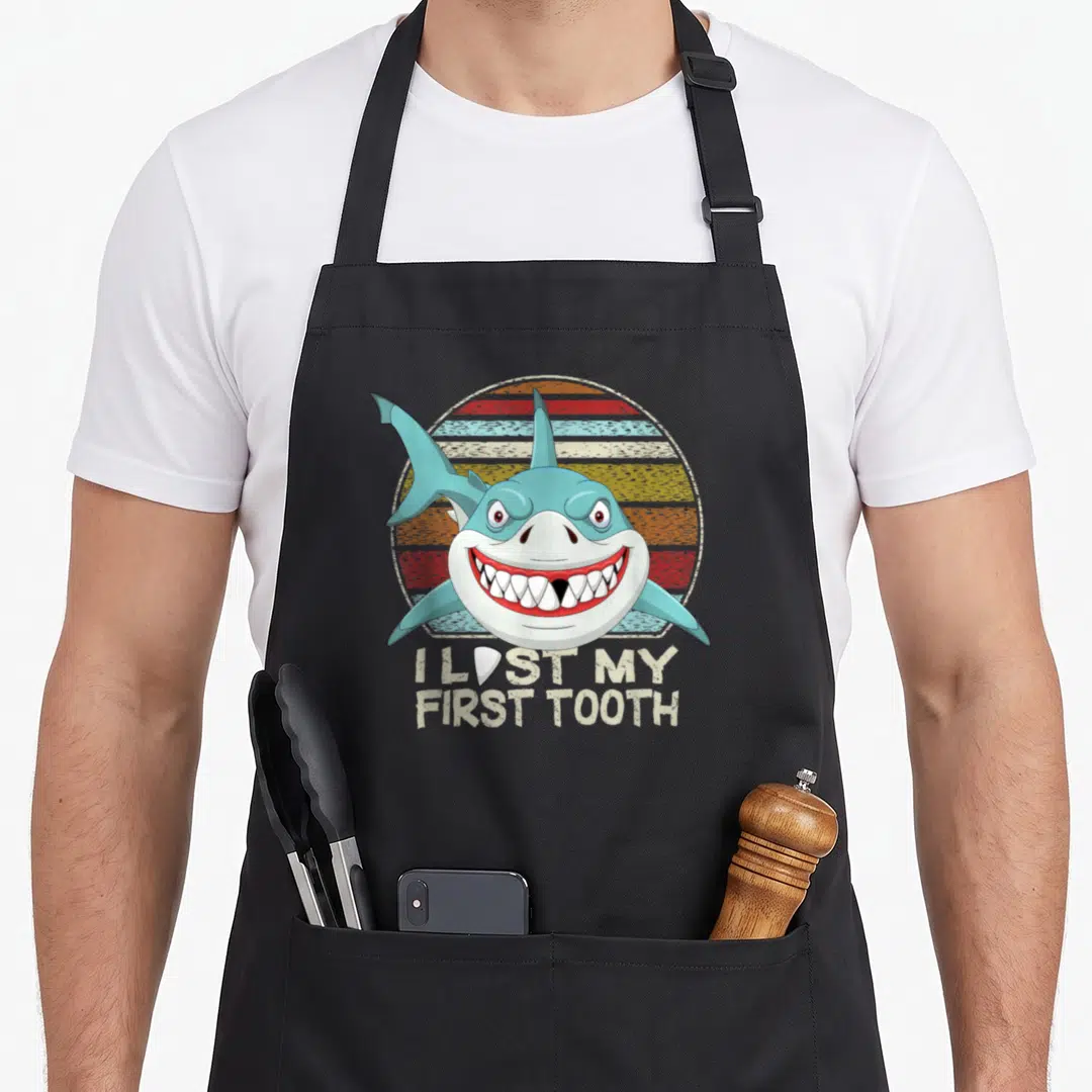 Fairy tooth apron - Cartoon shark head over retro stripes with I Lost My First Tooth text and tooth replacing letter O — shown on a black cotton canvas apron against a white background