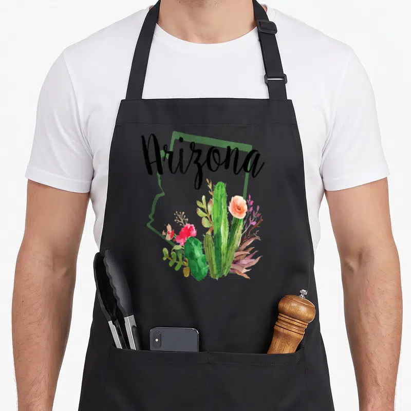 State cactus apron - Arizona text over green state outline with watercolor cacti and pink orange flowers — shown on a black cotton canvas apron against a white background
