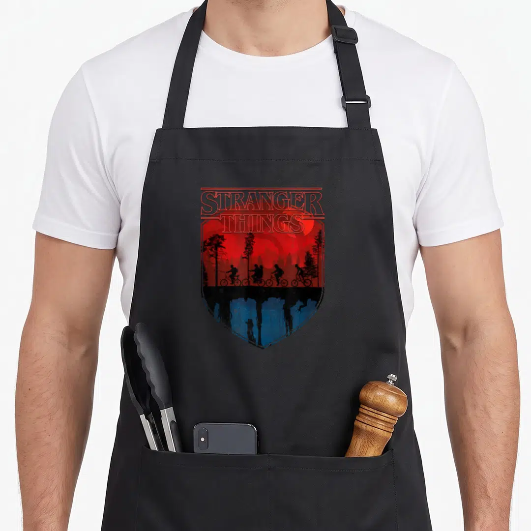 Shot trail apron - Shield graphic with STRANGER THINGS text, red sky, biking silhouettes, and blue underworld claw — shown on a black cotton canvas apron against a white background