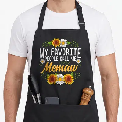 Call memaw mother's day aprons - Sunflowers and daisies framing My Favorite People Call Me Memaw text — shown on a black cotton canvas apron against a white background