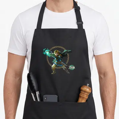Epic legend apron - Link casting cyan magic surrounded by a gold circle with game logo in corner — shown on a black cotton canvas apron against a white background